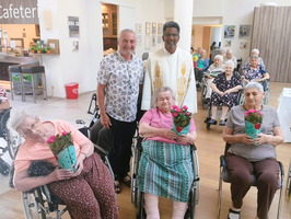 Eine Gruppe von Senioren in Rollstühlen posiert in einem Raum. Zwei Männer stehen in der Mitte, einer im weißen Gewand. Einige Senioren halten Blumensträuße, während andere lächeln. Im Hintergrund sind weitere Personen und eine Cafeteria sichtbar.