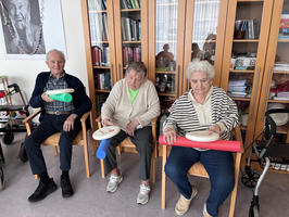 Drei Senioren sitzen auf Stühlen und halten Übungsgeräte in der Hand.