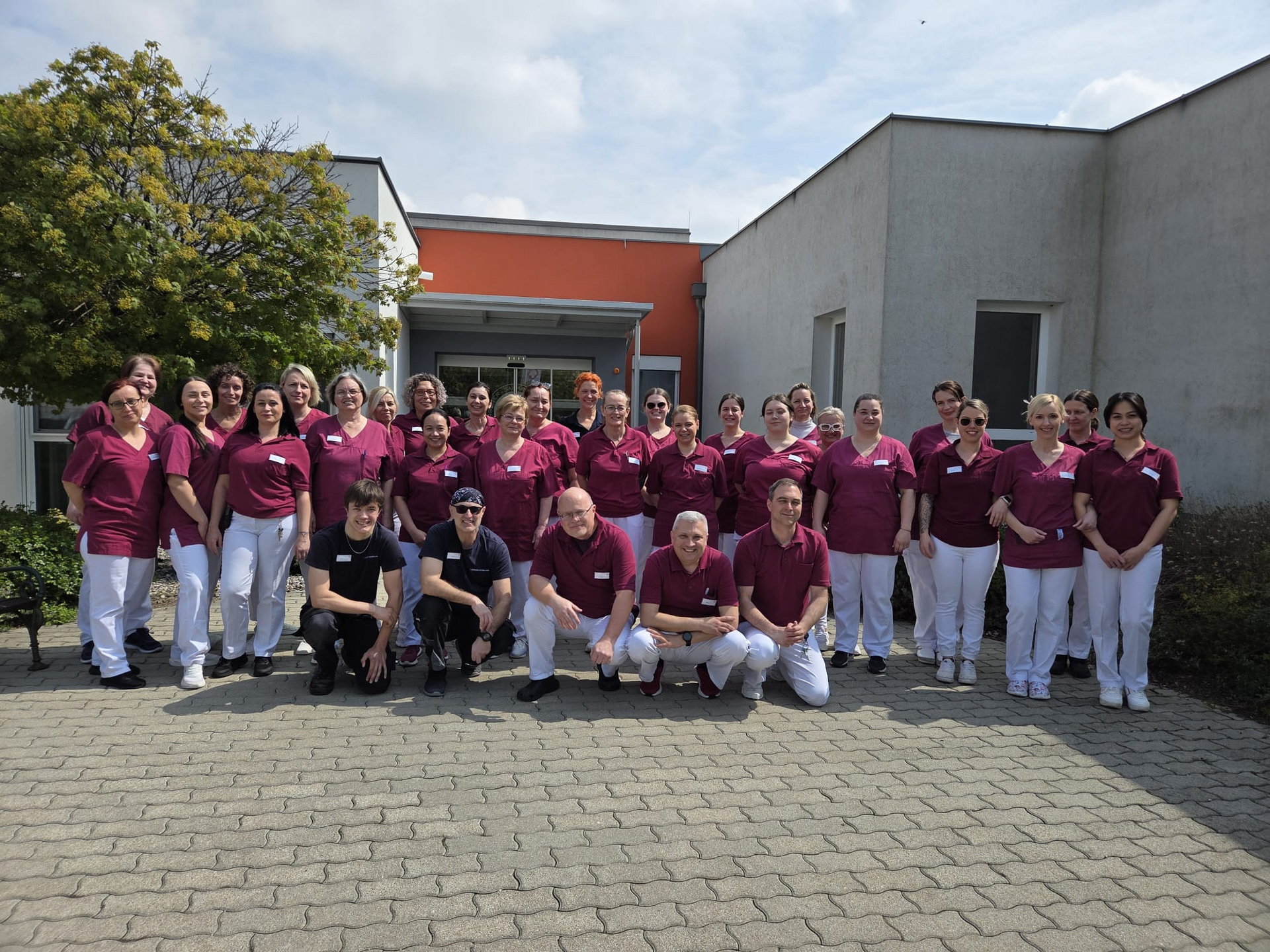 Eine Gruppe von etwa 40 Personen in medizinischer Kleidung steht auf einem Gehweg vor einem Gebäude. Sie lächeln in die Kamera, einige posieren. Im Hintergrund sind Bäume und das Gebäude sichtbar.