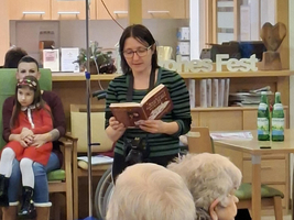 Frau sitzt und liest aus einem Buch, während Publikum aufmerksam zuhört.