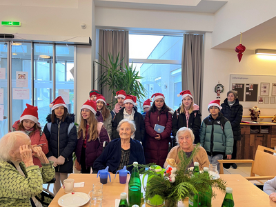 Eine Gruppe von Jugendlichen mit Weihnachtsmützen besucht ältere Menschen in einem Gemeinschaftsraum.