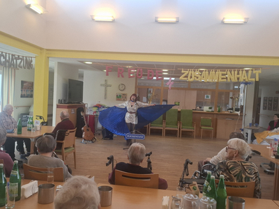 Eine Person mit einem großen blauen Umhang führt auf einer Bühne auf. Im Hintergrund stehen Stühle und Tische mit Gästen, die aufmerksam zuschauen. An der Wand hängen farbige Buchstaben, die 