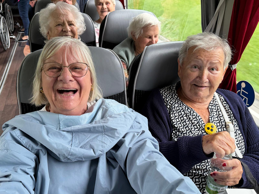 Eine Gruppe älterer Frauen sitzt in einem Bus. Im Vordergrund lächeln zwei Frauen fröhlich in die Kamera. Eine Frau links trägt eine Brille und eine graue Jacke, während die andere einen blauen Pullover und eine gelbe Brosche hat. Im Hintergrund sind weitere Frauen sichtbar.