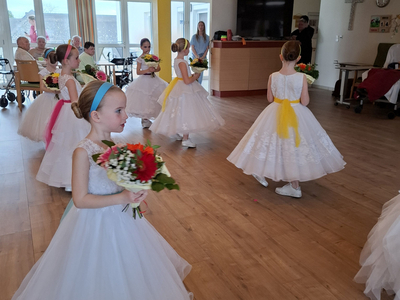 Mädchen in weißen Kleidern mit Blumensträußen bei einer Aufführung in einem Raum.