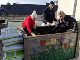 Drei Personen arbeiten gemeinsam an einem bunten Hochbeet. Sie füllen Erde ein, während mehrere Säcke mit Blumentopferde daneben stehen. Der Himmel ist blau und es ist sonnig.