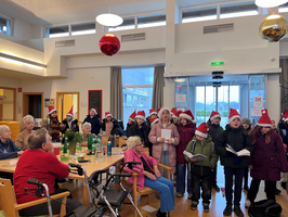 Kinder singen Weihnachtslieder im Seniorenheim, alle tragen Weihnachtsmützen.