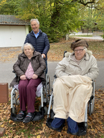 Ein Mann begleitet zwei ältere Frauen in Rollstühlen im Freien.