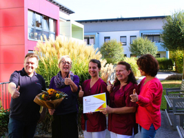 Fünf Personen halten ein Zertifikat und Blumenstrauß, alle zeigen Daumen hoch.