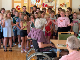 Eine Gruppe von Kindern steht in einem Raum und singt. Im Vordergrund sitzen ältere Personen, eine im Rollstuhl. Die Kinder wirken fröhlich und engagiert, während sie vor der kleinen Versammlung auftreten. Der Raum ist hell und hat eine freundliche Atmosphäre.