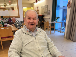 Ein lächelnder älterer Mann sitzt mit Essen am Tisch in einem Gemeinschaftsraum.