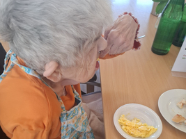 Eine ältere Frau mit grauen Haaren sitzt in einem Rollstuhl an einem Tisch. Sie trägt ein orangefarbenes Oberteil und eine Schürze. Auf dem Tisch befindet sich ein Teller mit Rührei und etwas Brot. Die Frau hält eine Hand an ihr Gesicht.