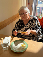 Eine ältere Frau mit grauen Haaren sitzt an einem Tisch und hält ein Stück Brot in der Hand. Vor ihr auf dem Tisch liegt ein leerer Teller mit zwei kleinen Schalen, die möglicherweise Öl oder Dip enthalten. Ein Glas Wasser steht ebenfalls auf dem Tisch. Tageslicht fällt durch ein Fenster.
