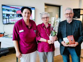 Drei Personen stehen zusammen mit Blumen und Geschenken in einem Empfangsraum.
