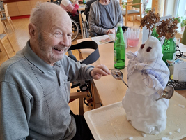 Ein älterer Mann lächelt zufrieden neben einem Schneemann mit Löffeln.