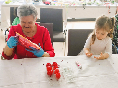 Eine ältere Frau in rotem Oberteil und blauen Handschuhen hält einen Beutel mit roter Farbschicht und lächelt, während ein kleines Mädchen in einem hellen Oberteil an einem Tisch sitzt und mit den Händen spielt. Im Vordergrund sind einige rote Ostereier zu sehen.