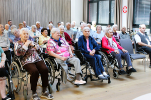 Eine Gruppe älterer Menschen, viele in Rollstühlen, sitzen in einem Raum.