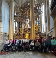 Eine Gruppe von etwa 30 Personen, hauptsächlich in Rollstühlen, steht in einem Innenraum mit einer kunstvollen barocken Altaranlage. Die Wände sind hoch und mit großen Fenstern versehen, die viel Licht hereinlassen. Die Menschen lächeln und scheinen an einem Ausflug teilzunehmen.