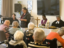 Gottesdienst mit älteren Menschen, einer Person liest am Mikrofon vor.