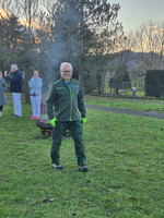 Eine Person in sportlicher, grüner Kleidung steht auf einer Wiese, im Hintergrund sind weitere Menschen zu sehen, einige sprechen miteinander. Es ist ein sonniger Abend, und Rauch steigt von einem Grill auf.