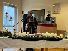 Ein Priester hält eine Ansprache vor einem Tisch mit Kerzen und Blumen.