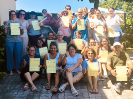 Eine Gruppe von 22 Menschen sitzt und steht im Freien und hält stolz gelbe Zertifikate in den Händen. Sie lächeln und zeigen Daumen nach oben. Die Atmosphäre wirkt festlich und zufrieden. Im Hintergrund sind Bäume und ein sonniger Himmel sichtbar.