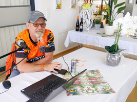 Ein Mann mit Brille und Bart sitzt an einem Tisch mit einem Laptop. Er trägt ein orangefarbenes T-Shirt und hat ein Mikrofon neben sich. Auf dem Tisch stehen eine Getränkedose, ein Notizblock und eine Topfpflanze. Im Hintergrund sind Flaschen und Dekorationen sichtbar.