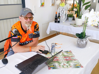 Ein Mann mit Brille und Bart sitzt an einem Tisch mit einem Laptop. Er trägt ein orangefarbenes T-Shirt und hat ein Mikrofon neben sich. Auf dem Tisch stehen eine Getränkedose, ein Notizblock und eine Topfpflanze. Im Hintergrund sind Flaschen und Dekorationen sichtbar.