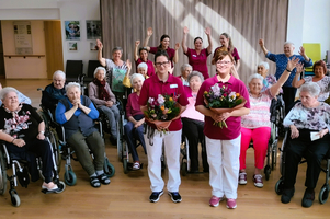 Gruppenszene mit Senioren und Pflegekräften, die Blumensträuße halten und jubeln.