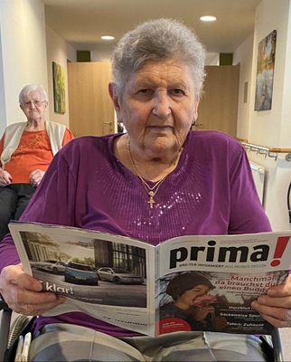 Eine ältere Frau sitzt im Rollstuhl und liest eine Zeitschrift.