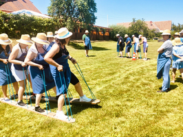 Eine Gruppe von Menschen in Sommerkleidung und Strohhüten steht auf einer Wiese. Einige von ihnen balancieren auf einem langen Holzbrett und verwenden Seile, um gemeinsam zu arbeiten. Im Hintergrund beobachten andere Teilnehmer das Geschehen.