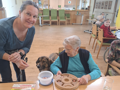 Eine Betreuerin interagiert mit einem Hund vor einer Gruppe älterer Menschen.