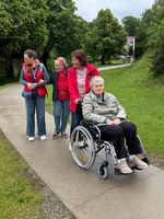 Vier Frauen stehen auf einem Gehweg in einem Park. Eine Frau sitzt im Rollstuhl, während die anderen um sie stehen. Sie scheinen miteinander zu sprechen und genießen die frische Luft an einem bewölkten Tag. Grüne Bäume und Rasen umgeben die Szene.