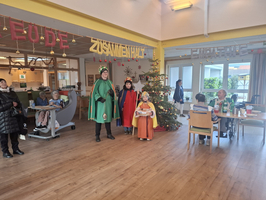 Drei verkleidete Personen stehen vor einem geschmückten Weihnachtsbaum in einem Gemeinschaftsraum.