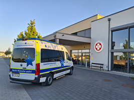 Ein Lieferwagen mit buntem Design steht vor dem Eingangsbereich des Kompetenzzentrums. Die Fassade des Gebäudes ist modern, mit großen Fenstern und einem sichtbaren Logo. Im Hintergrund sind Bäume und eine klare Himmel zu sehen.