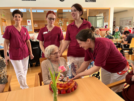 Eine ältere Frau sitzt an einem Tisch und lächelt, während Pflegekräfte um sie versammelt sind. Vor ihr steht ein Geburtstagskuchen mit Früchten, und eine der Pflegekräfte überreicht ihr ein Geschenk. Die Atmosphäre ist festlich und fröhlich.