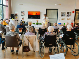 Eine Gruppe älterer Menschen in Rollstühlen sitzt in einem Raum. Vor ihnen steht eine Person in liturgischer Kleidung, die eine Zeremonie leitet. Im Hintergrund spielt ein Klavier, und an der Wand ist ein Bild mit zwei Kreuzen zu sehen.