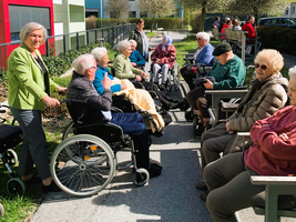 Gruppenszene im Freien mit älteren Menschen in Rollstühlen und auf Bänken. Sie sind in entspannter Atmosphäre zusammen, genießen die Sonne. Eine Person steht in der Nähe. Im Hintergrund sind weitere Menschen und Pflanzen sichtbar.