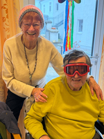 Eine fröhliche Frau steht hinter einem Mann in einer Sitzschale mit roten Brillen.