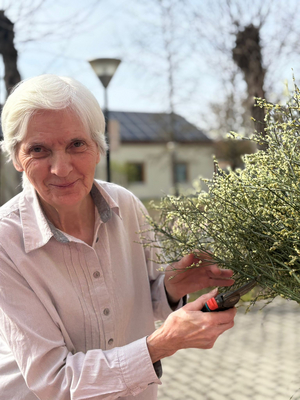 Eine ältere Frau mit grauen Haaren lächelt und hält einen Ast mit kleinen, weißen Blüten in einer Hand. In der anderen Hand hält sie eine Gartenschere. Im Hintergrund sind Bäume und ein Gebäude sichtbar. Es ist ein sonniger Tag.