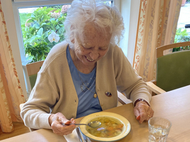 Eine ältere Frau lächelt beim Essen von Suppe an einem Tisch.