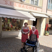 Zwei Frauen stehen vor einem Geschäft mit dem Namen „S.E.E.B.A.C.H.E.R“. Eine Frau trägt eine Schürze und die andere sitzt im Rollstuhl. Im Hintergrund sind Fenster mit verschiedenen Produkten und Dekorationen zu sehen.
