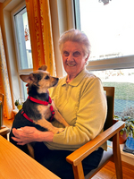 Eine ältere Frau sitzt an einem Tisch und hält einen kleinen Hund auf ihrem Schoß. Beide schauen sich an und lächeln. Im Hintergrund sind große Fenster mit Tageslicht und einige Pflanzen zu sehen.