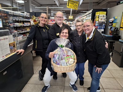 Fünf Personen stehen in einem Supermarkt zusammen. Eine Frau hält einen geflochtenen Korb mit verschiedenen Produkten. Die Gruppe lächelt und posiert freundlich vor der Kasse. Im Hintergrund sind Regale mit Waren sichtbar.