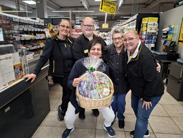 Fünf Personen stehen in einem Supermarkt zusammen. Eine Frau hält einen geflochtenen Korb mit verschiedenen Produkten. Die Gruppe lächelt und posiert freundlich vor der Kasse. Im Hintergrund sind Regale mit Waren sichtbar.