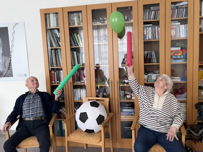 Zwei Senioren heben bunte Stäbe und einen Ball in einer Einrichtung.