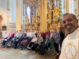 Eine Gruppe von Senioren in Rollstühlen posiert lächelnd in einer Kirche. Ein Pfarrer steht vorne rechts und macht ein Selfie. Im Hintergrund sind kunstvolle barocke Skulpturen und Säulen zu sehen. Das Licht fällt durch große Fenster auf die Szene.