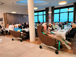 Ältere Menschen sitzen in einem Raum mit Tischen und Stühlen.