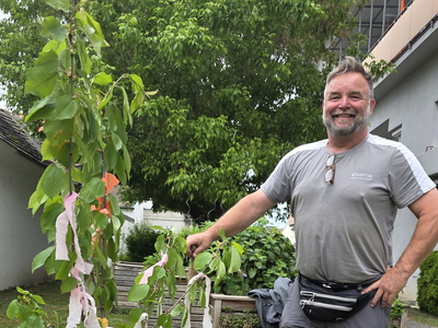 Ein lächelnder Mann steht neben einem kleinen Baum, der mit bunten Bändern geschmückt ist. Er hält eine Gartengabel in der Hand und trägt eine graue T-Shirt und graue Hose. Im Hintergrund sind eine grüne Wiese und einige Pflanzen zu sehen.