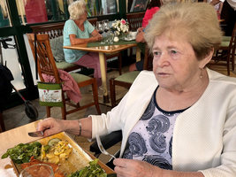 Eine ältere Frau sitzt an einem Tisch mit einem Teller Essen und einem Bier.