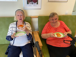 Zwei ältere Frauen sitzen auf einer Bank und halten Essen auf Tellern.